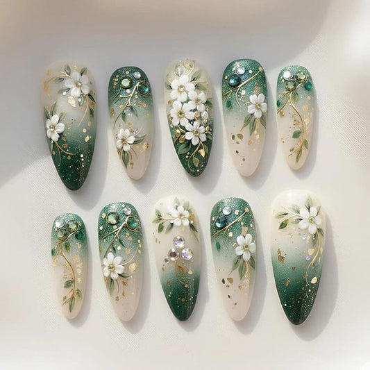 Ten almond-shaped press on nails displayed in two rows with a green gradient base, hand-painted white florals, gold leaf foil accents, and crystal rhinestone embellishments — perfect for wedding, bridal, or cottagecore aesthetic looks.