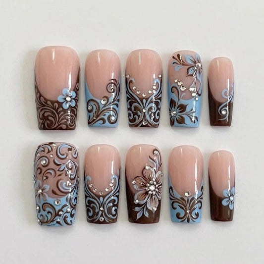 A detailed macro shot of brown and blue swirl nail art on luxury gel fake nails, showcasing professional-grade hand-painted patterns and sparkling crystal embellishments.