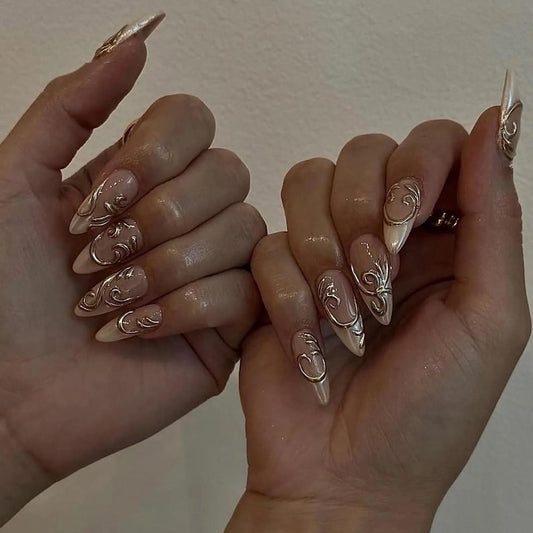 Shopify product display of long almond-stiletto false nails showcasing a semi-sheer nude base coat decorated with sculpted 3D gold metal patterns and French chrome tips.