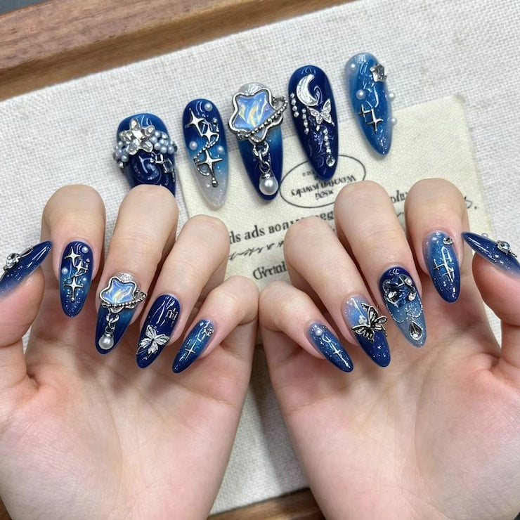 Close-up of 3D butterfly and crescent moon nail art designs on almond-shaped press-on nails in royal blue and silver glitter gradients.