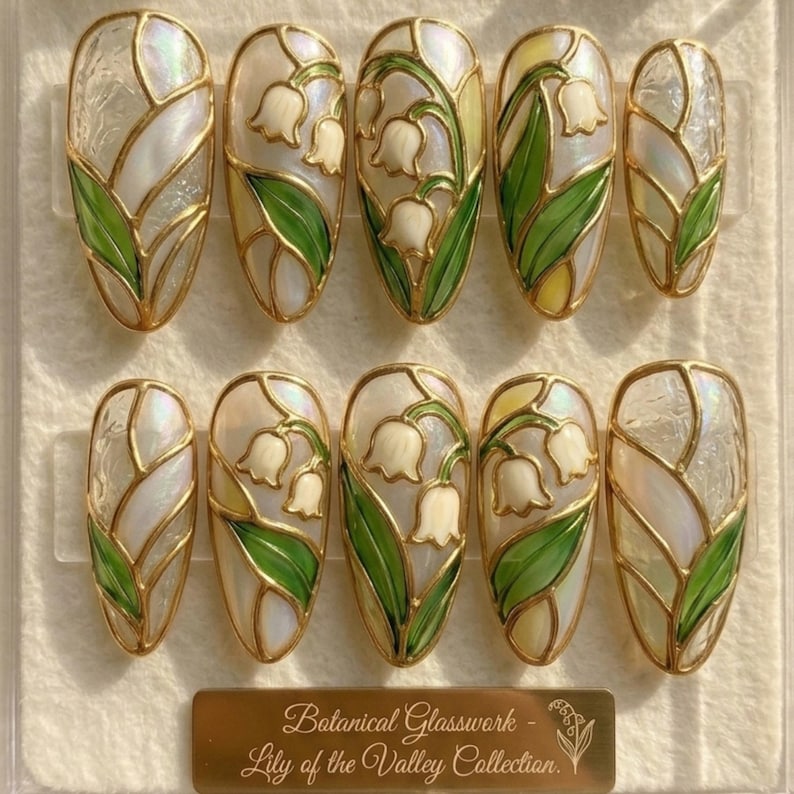 Two rows of ten custom almond press-on nails from JRNAIL featuring an iridescent pearl base decorated with 3D sculpted gold borders and hand-painted green and white lily of the valley flowers.