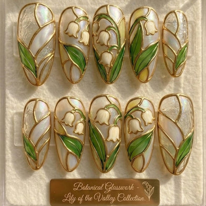 Two rows of ten custom almond press-on nails from JRNAIL featuring an iridescent pearl base decorated with 3D sculpted gold borders and hand-painted green and white lily of the valley flowers.