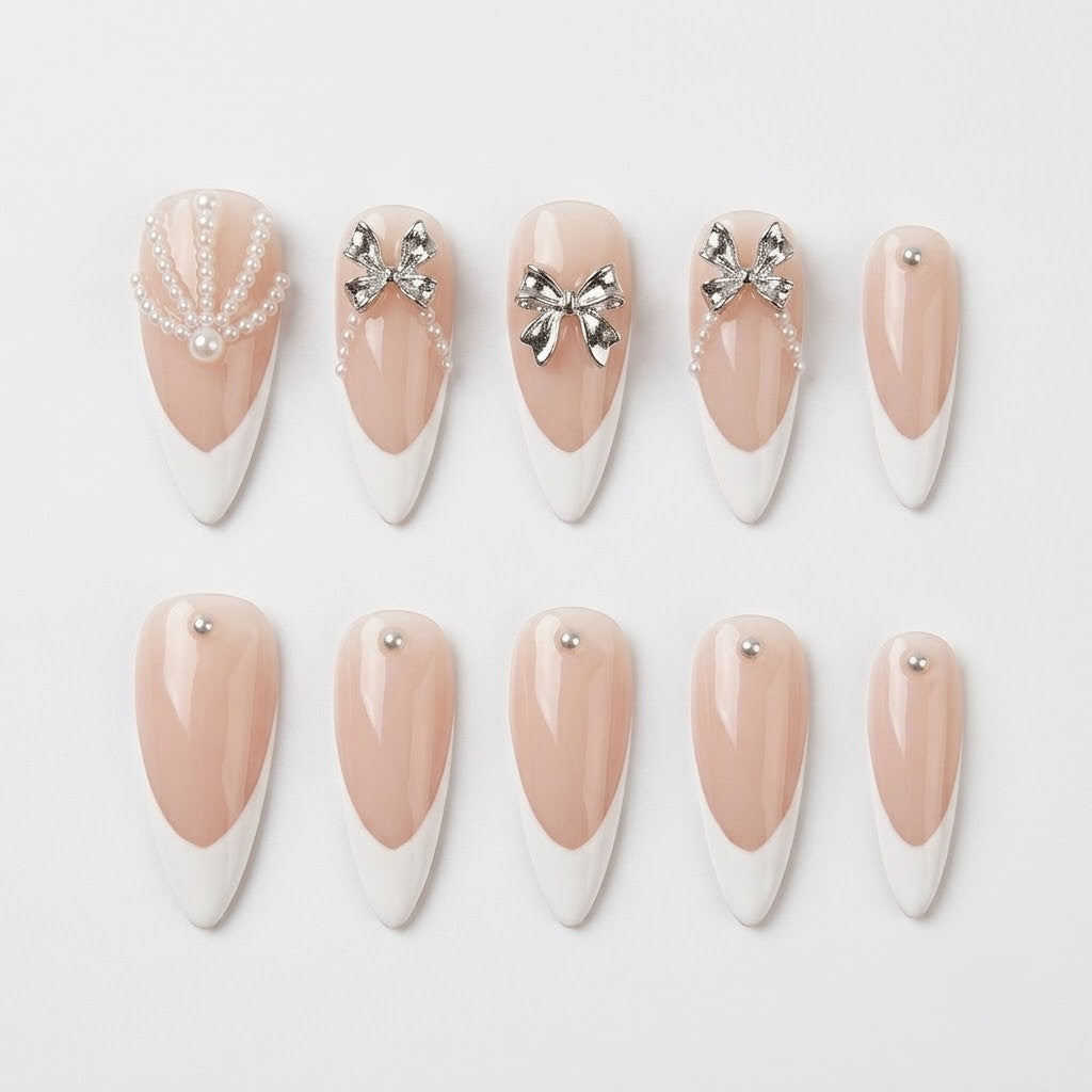 Close-up of handmade false nails displaying a high-gloss sheer nude base, sharp white French V-cut tips, raised sculptural silver bows, and delicate miniature pearl string accents.