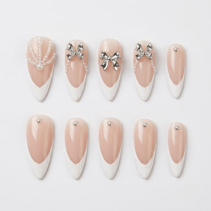 Close-up of handmade false nails displaying a high-gloss sheer nude base, sharp white French V-cut tips, raised sculptural silver bows, and delicate miniature pearl string accents.