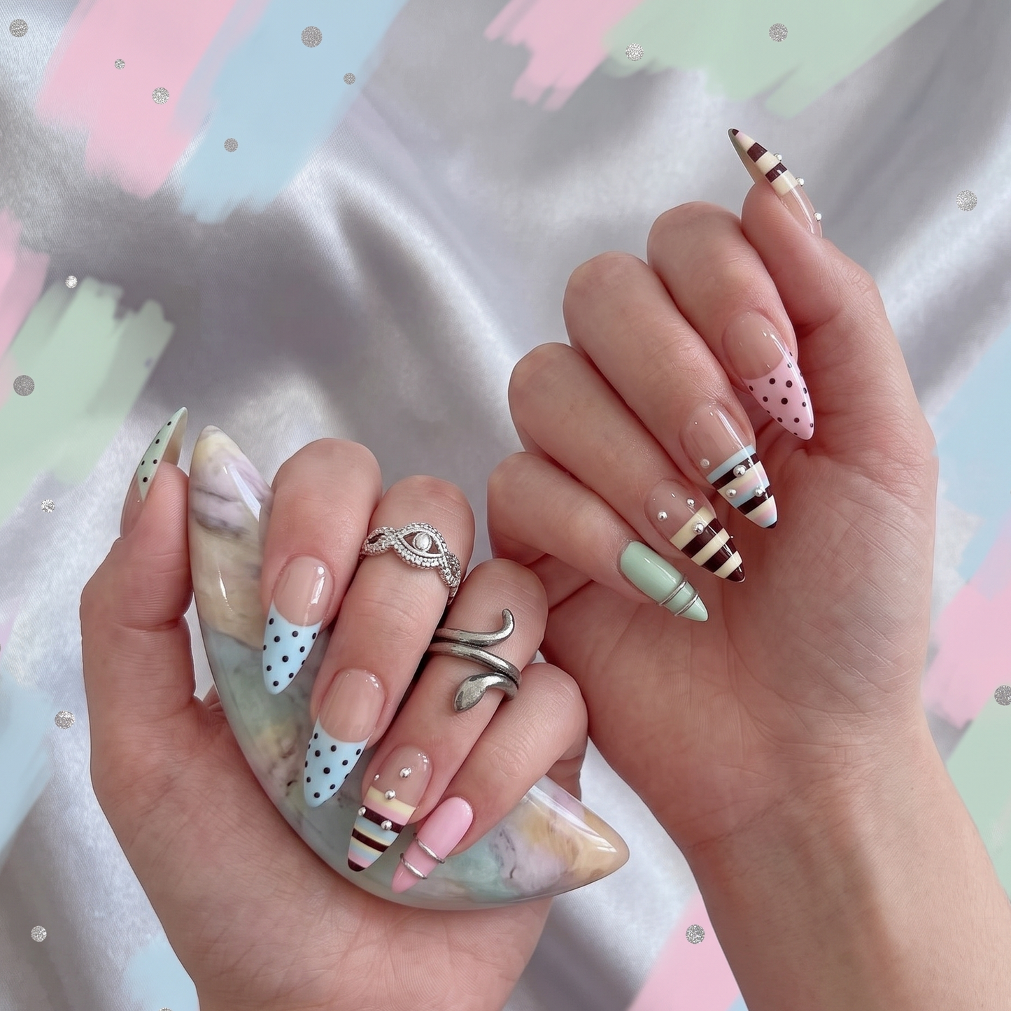 A pair of hands showcasing a custom luxury press-on nail set featuring pastel striped and polka dot French tips, accented by silver rhinestones and textured metallic rings.