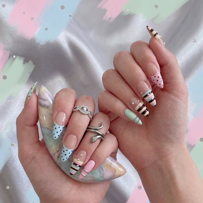 A pair of hands showcasing a custom luxury press-on nail set featuring pastel striped and polka dot French tips, accented by silver rhinestones and textured metallic rings.