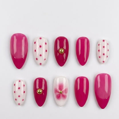 A full 10-piece set of handcrafted almond-shaped press-on nails featuring glossy hot pink, pink polka dots, and a 3D blush orchid flower with gold bead accents, arranged neatly on a clean white background.