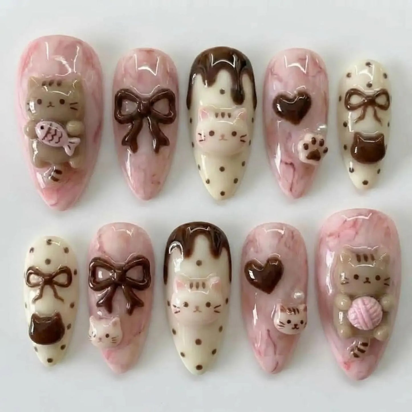 Elegant almond-shaped reusable false nails adorned with intricate 3D kitty paws, yarn balls, and dark chocolate hearts, displayed beautifully against a coordinating pastel pink background.