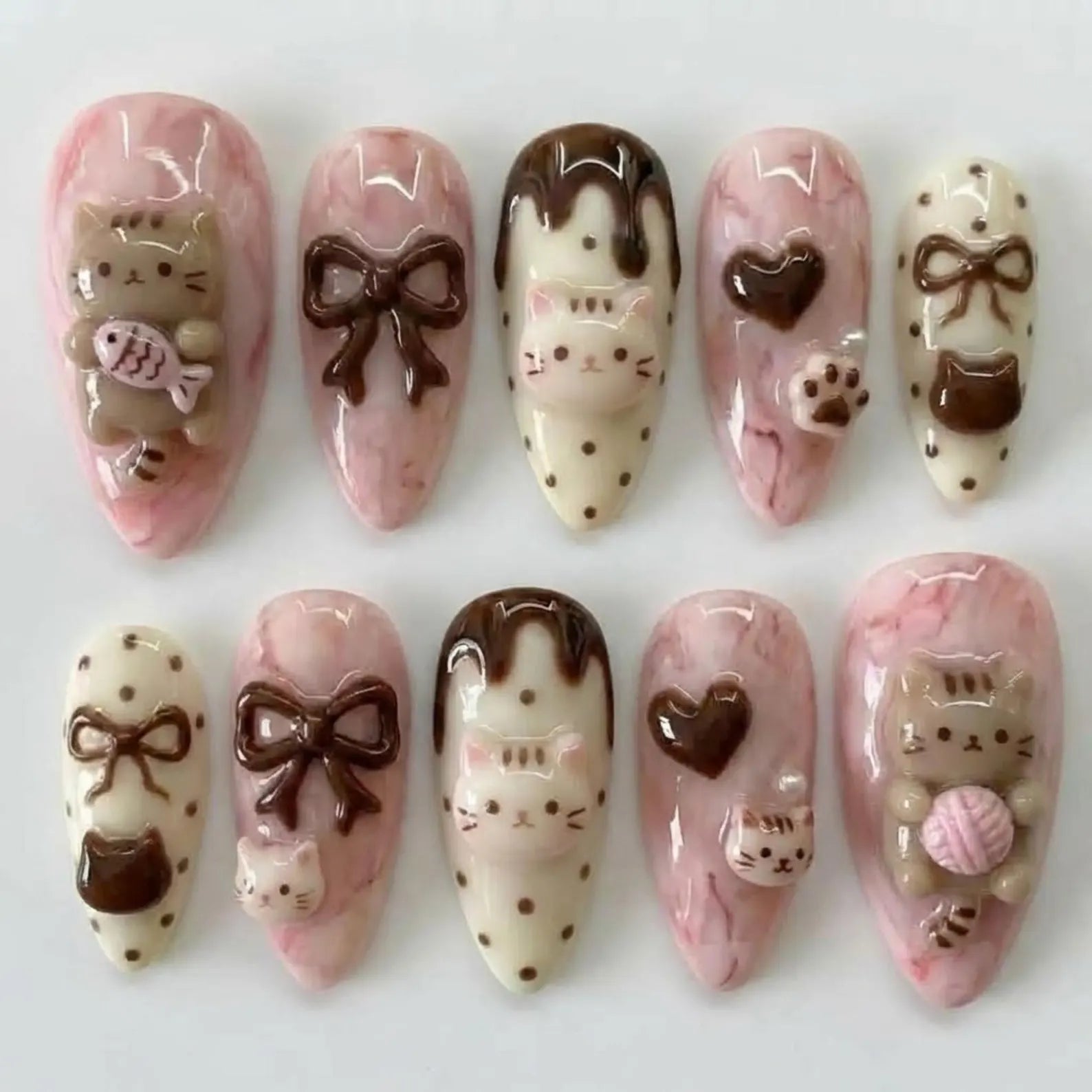 Elegant almond-shaped reusable false nails adorned with intricate 3D kitty paws, yarn balls, and dark chocolate hearts, displayed beautifully against a coordinating pastel pink background.