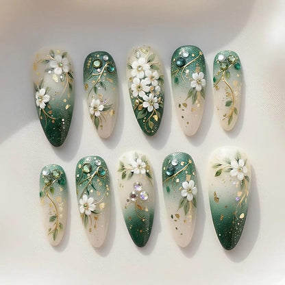 Ten almond-shaped press on nails displayed in two rows with a green gradient base, hand-painted white florals, gold leaf foil accents, and crystal rhinestone embellishments — perfect for wedding, bridal, or cottagecore aesthetic looks.