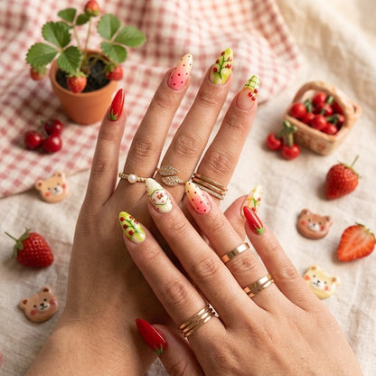 A close-up view of two hands modeling almond-shaped press-on nails featuring a vibrant red strawberry theme, green plaid patterns, and gold rings against a cozy cottagecore background with fresh strawberries.