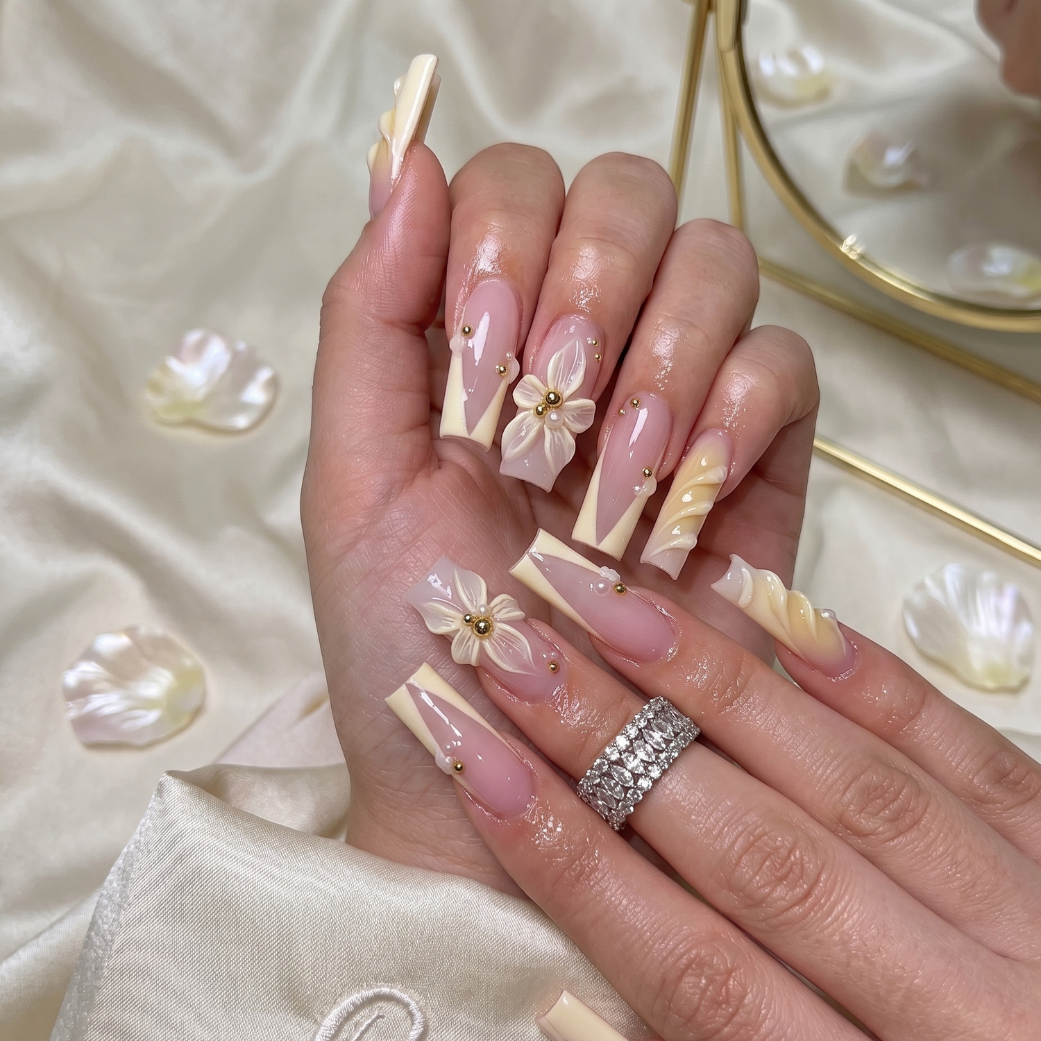 A display of ten long, coffin-shaped press-on nails from the Ethereal Blooms collection, arranged on a textured cream paper. This unique design features a mix of intricate hand-sculpted 3D flowers with gold beads, elegant pink and cream geometric French tips, and a textured white accent nail.