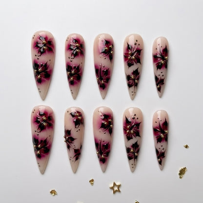 Close-up of premium handmade false nails displaying a soft ivory base, deep plum airbrushed floral accents, delicate black speckles, and embedded gold metallic beads.
