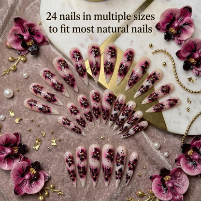 A full 24-piece luxury press-on nail set featuring sharp stiletto tips with hand-painted burgundy orchid flower designs and gold caviar beads, arranged in an elegant fan layout on a velvet and marble background with text reading '24 nails in multiple sizes to fit most natural nails.
