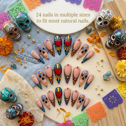 A premium 24-piece luxury press-on nail set featuring dramatic stiletto tips hand-painted with colorful sugar skulls, neon sunburst patterns, and red hearts, laid out flat amidst Day of the Dead decor, marigolds, and colorful banners with text reading '24 nails in multiple sizes to fit most natural nails.