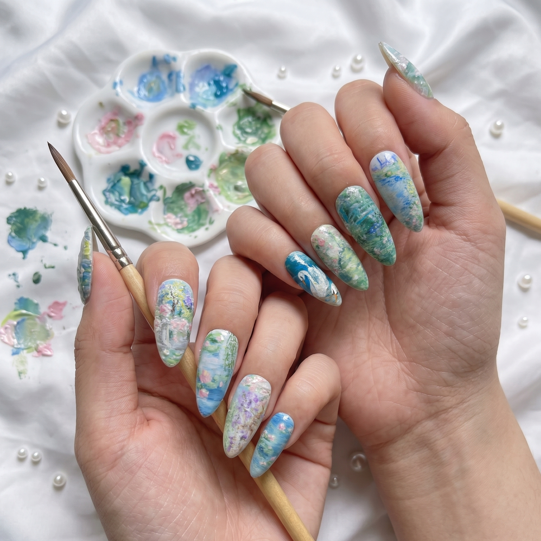 A complete set of ten stiletto-shaped press-on nails featuring hand-painted Impressionist art inspired by Monet's gardens, including water lilies, roses, and weeping willows, neatly arranged in two rows on a clean white background with scattered pearls.
