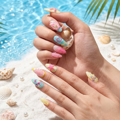Close-up shot of a hand wearing JRNAIL tropical 3D press-on nails with starfish, hibiscus flowers, and bikini designs over a sandy beach and blue water background