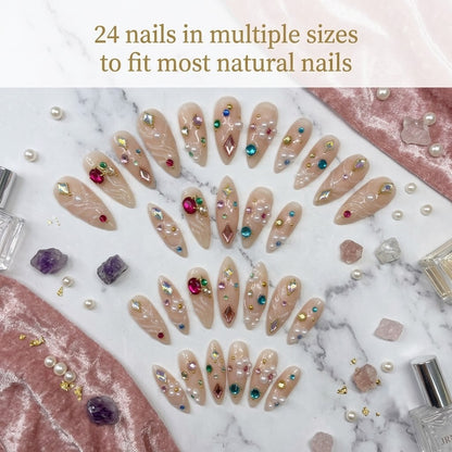 A premium 24-piece luxury press-on nail set featuring sharp stiletto tips with a sheer jelly nude base, clear 3D fluid ripples, and multi-colored gemstones, arranged beautifully in rows over a white marble surface accented with pink velvet cloth, raw crystals, and pearls under a text banner reading '24 nails in multiple sizes to fit most natural nails.