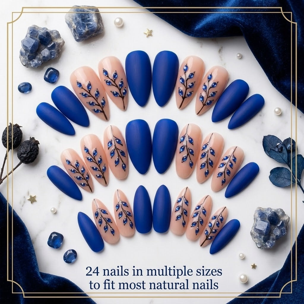 A premium 24-piece luxury press-on nail set featuring sharp stiletto tips with a mix of velvety matte royal blue finishes and glossy nude accent nails decorated with hand-painted black branches and 3D metallic blue leaf jewels, arranged neatly in rows over a white marble surface accented with deep blue velvet fabric, sapphire gemstones, and text reading '24 nails in multiple sizes to fit most natural nails.