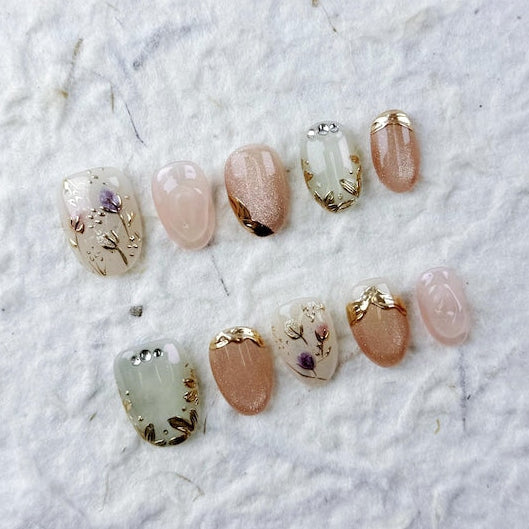 Luxury reusable gel false nails showcasing a mix of neutral milky bases with 3D botanical nail art and textured gold borders.