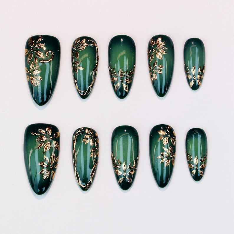 A luxury set of stiletto-shaped reusable false nails in deep jade green, decorated with intricate metallic gold leaf patterns, 3D floral accents, and a glossy gel topcoat, showcased for an online boutique
