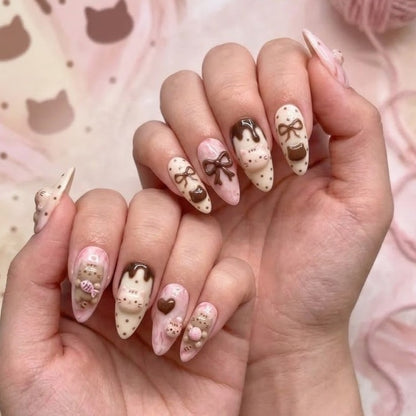 A pair of hands showcasing a custom luxury press-on nail set featuring 3D sculpted brown tabby cats, chocolate bows, and glossy drizzle patterns over a soft pink marbled base.