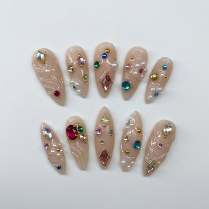 Close-up of handmade maximalist false nails showcasing dimensional clear gel swirls, vibrant sapphire blue, ruby red, and emerald green rhinestones, alongside miniature pearl clusters and gold beads.
