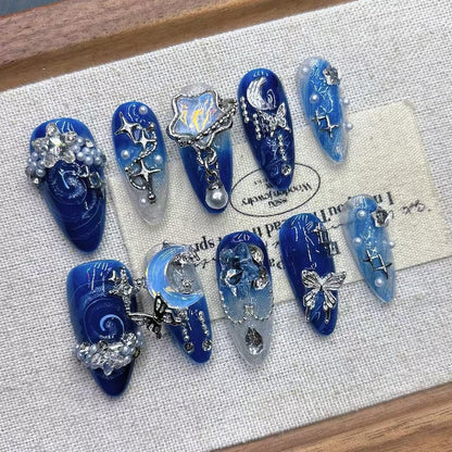 Flatlay display of starry night fake nails by JRNAIL, showcasing deep navy blue hues, star rhinestones, and silver chain accents on a wooden-framed canvas.
