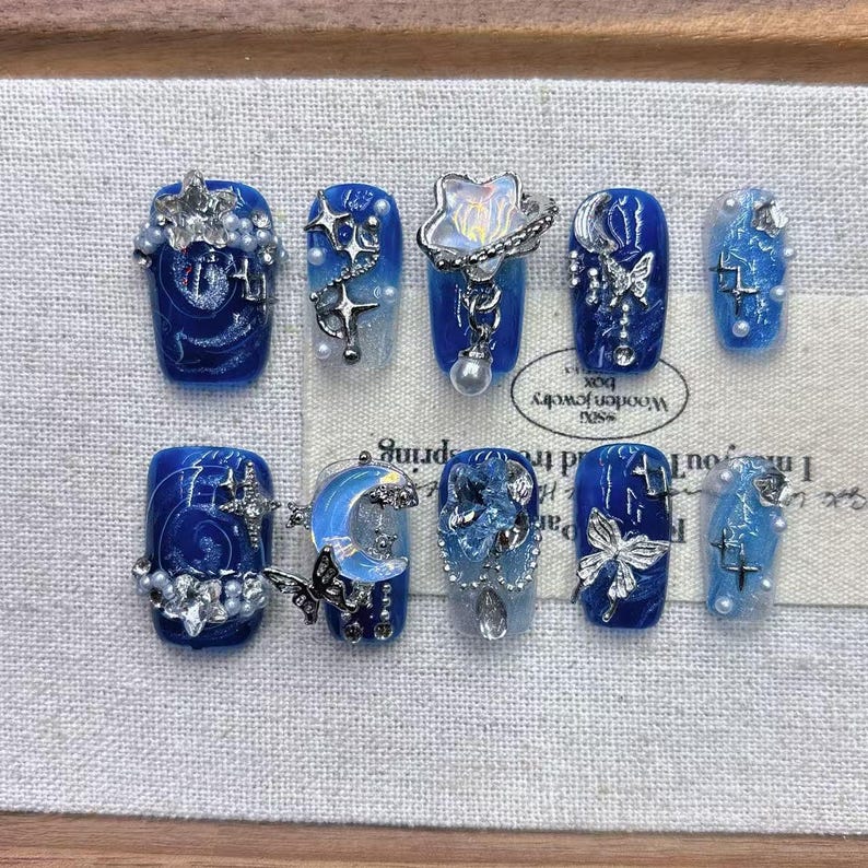 Luxury glue-on nails with a galaxy theme, showing intricate hand-painted blue swirls, embedded pearls, and dangling metallic silver star charms.
