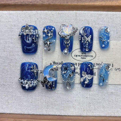 Luxury glue-on nails with a galaxy theme, showing intricate hand-painted blue swirls, embedded pearls, and dangling metallic silver star charms.