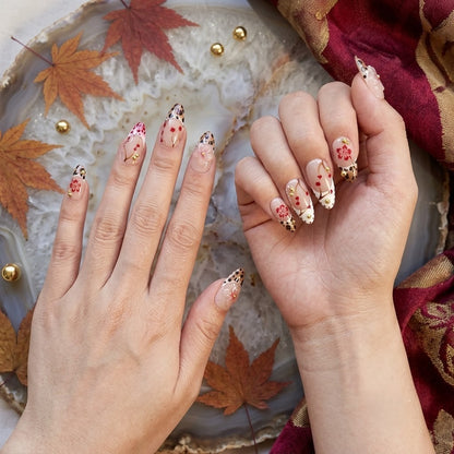 A close-up view of two hands modeling JRNAIL's Autumn Maple and Leopard print almond press-on nails. The glossy, almond-shaped nails feature detailed red maple branches, leopard print tips, 3D acrylic flowers, and gold bead accents. The hands are posed elegantly over a crystal geode tray surrounded by red autumn leaves and luxury burgundy silk.