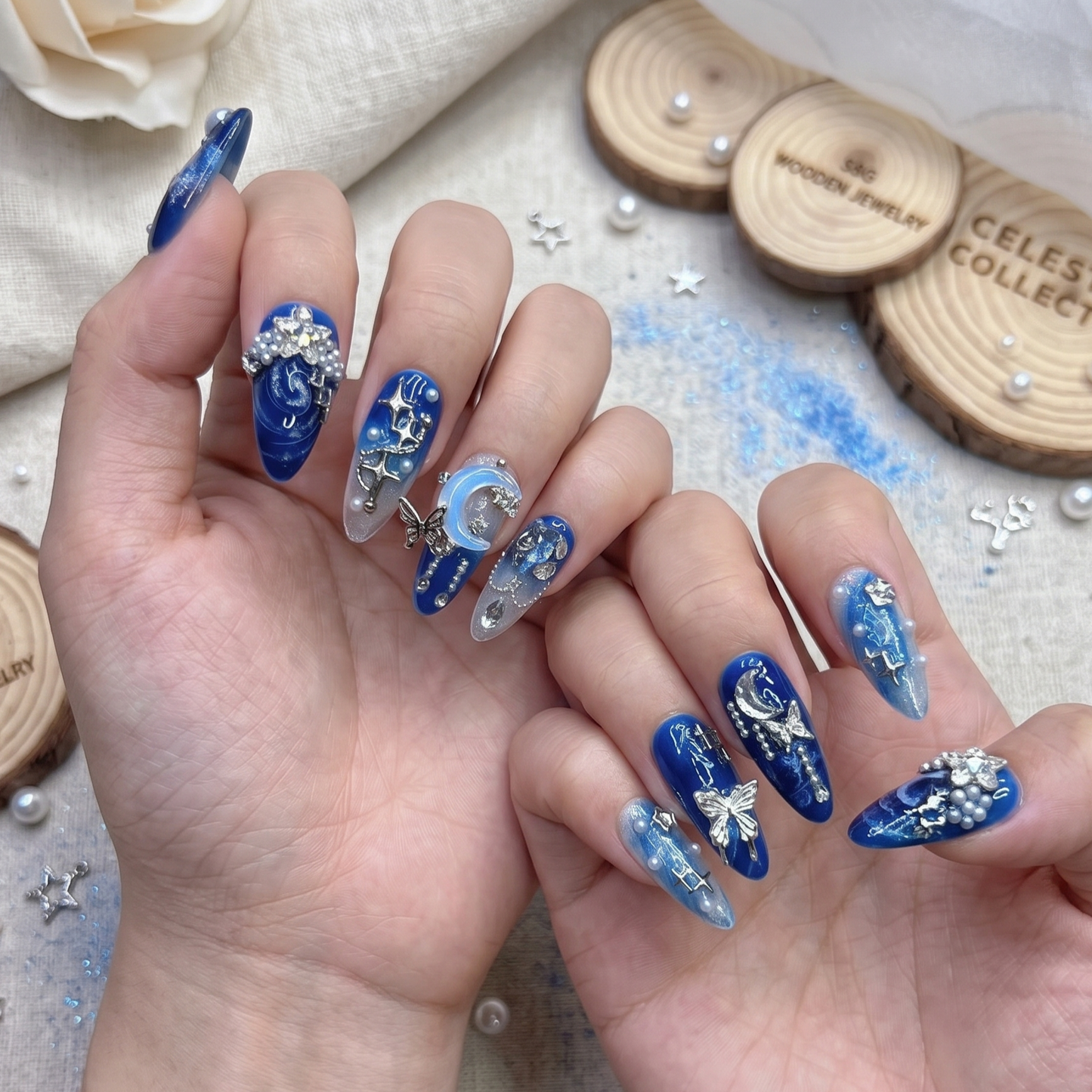 A set of 10 handcrafted celestial press-on nails displayed on a beige canvas background, featuring deep blue gel polish, silver moons, 3D stars, and butterfly charms.