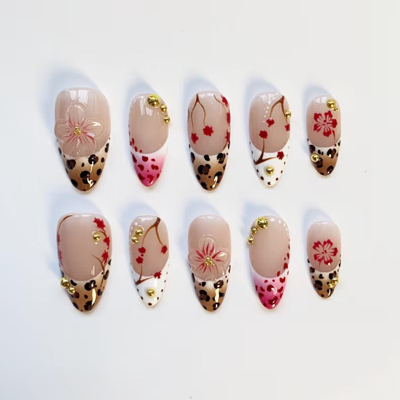 An overhead flat lay showing 10 individual almond press-on nails from JRNAIL arranged in two rows against a clean white background. The image clearly showcases the variety of patterns, including raised 3D pink flowers, leopard spots, painted red branches, and golden micro-stud embellishments on a nude base.