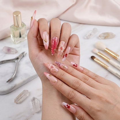An organized layout of luxury stiletto fake nails by JRNAIL displaying tropical floral patterns, vibrant animal prints, and gold metallic charms on a white textured surface with pink satin.