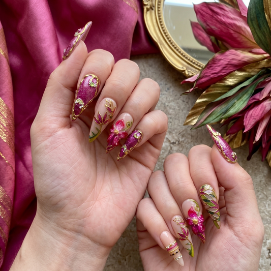 Hand model showcasing JRNAIL's magenta floral press-on nails with 3D hibiscus petals and gold leaf detailing against a rich pink satin background.