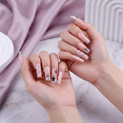 A close-up of a hand model showing two hands wearing JRNAIL long coffin fake nails with a nude base, black starburst graphics, and luxury crystal cross embellishments.
