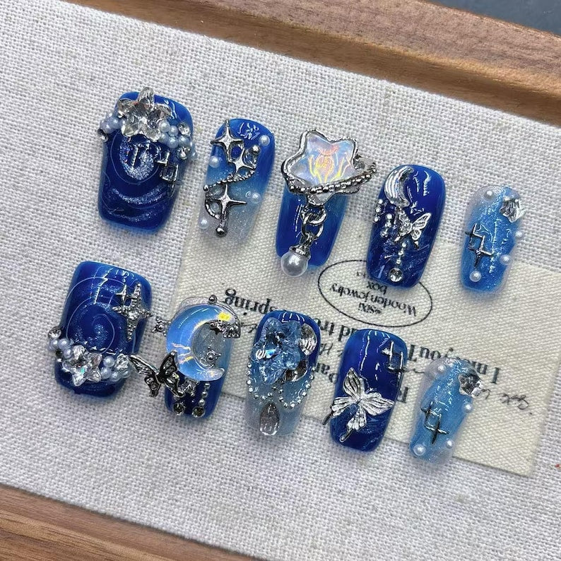 A flawless salon-quality manicure featuring luxury blue gel press-ons adorned with silver moon and butterfly charms, posed over wooden prop discs.